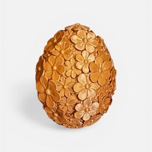 May include: A decorative, egg-shaped ornament in a golden hue. The surface is densely covered with three-dimensional flower designs of different sizes. The object is set against a plain white backdrop, emphasizing the detailed floral pattern.
