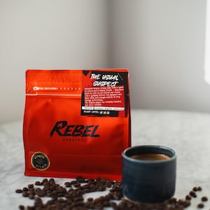 May include: A red coffee bag with the text "REBEL roastery" and "THE USUAL SUSPECT" next to a dark blue ceramic cup filled with coffee. Coffee beans are scattered around the cup and bag.