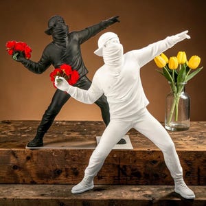 Op de afbeelding: Twee sculpturen van figuren in actie, één zwart en één wit. De zwarte figuur houdt rode rozen vast, de witte figuur heeft een boeket gele tulpen in een vaas in de buurt. Beide figuren dragen gezichtsbedekkingen en zijn in een werpbeweging.