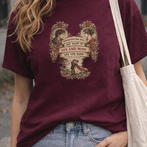 May include: Burgundy t-shirt featuring a romantic graphic with two portraits and the quote "WHATEVER OUR SOULS ARE MADE OF, HIS AND MINE ARE THE SAME." The design includes floral elements and a couple embracing. The shirt is paired with blue jeans.