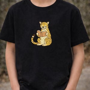 Può includere: T-shirt nera con un ghepardo dei cartoni animati che legge un libro. Il ghepardo è giallo con macchie nere e indossa gli occhiali. Il libro è marrone. La maglietta è indossata da un bambino.