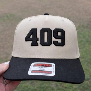May include: Tan and black baseball cap with the number "409" embroidered in black on the front. The cap has a black brim and a tag with the brand name "OTTO".