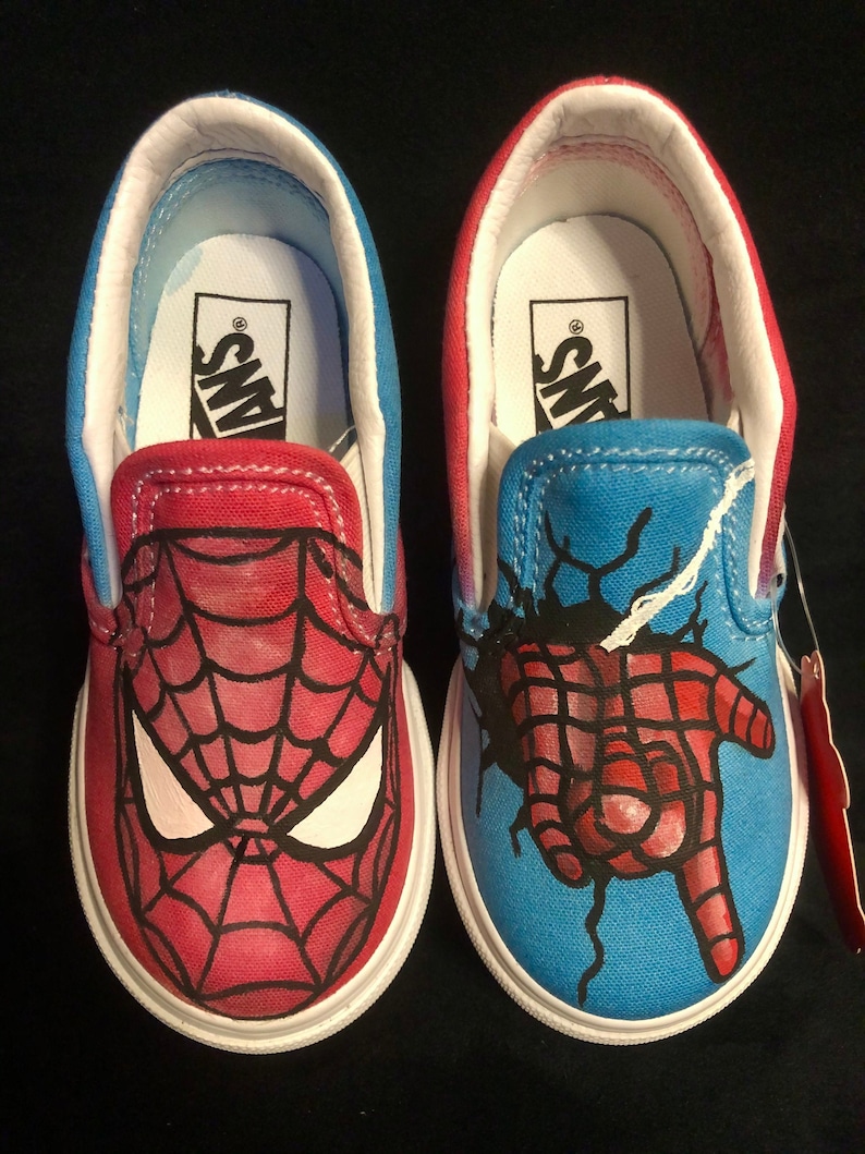 May include: A pair of red and blue Vans slip-on shoes with a custom hand-painted design featuring Spider-Man. One shoe has a close-up of Spider-Man's face, while the other shoe has a hand breaking through a wall with Spider-Man's web design.