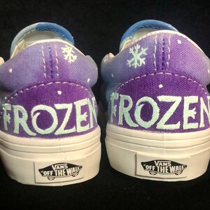 Puede incluir: Un par de zapatos Vans slip-on de color morado y azul con un dise&ntilde;o de copo de nieve blanco y la palabra "FROZEN" pintada en la parte posterior de cada zapato.