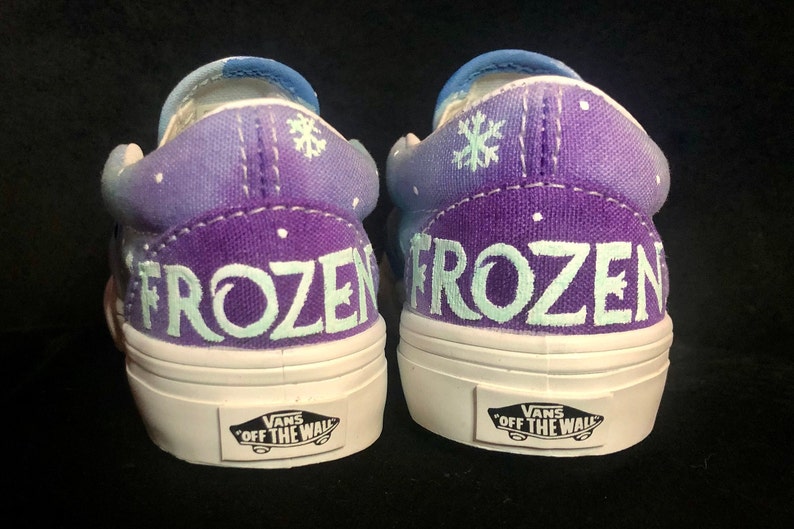 Puede incluir: Un par de zapatos Vans slip-on de color morado y azul con un dise&ntilde;o de copo de nieve blanco y la palabra "FROZEN" pintada en la parte posterior de cada zapato.