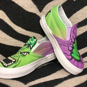 Może przedstawiać: Para zielono-fioletowych pł&oacute;ciennych but&oacute;w typu slip-on z nadrukiem kresk&oacute;wkowego potwora. Jedno z but&oacute;w przedstawia zieloną twarz potwora z szeroko otwartą paszczą i ostrymi zębami. Drugie z but&oacute;w przedstawia zieloną twarz potwora na fioletowym tle z czarnymi błyskawicami.