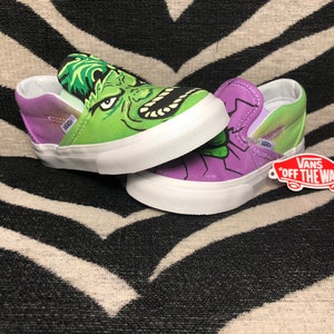 Może przedstawiać: Para zielono-fioletowych but&oacute;w Vans slip-on z grafiką Hulka na jednym bucie i fioletowo-zieloną grafiką na drugim. Buty mają białą podeszwę i metkę Vans "Off The Wall".