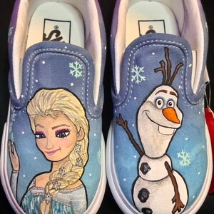 Puede incluir: Un par de zapatos de lona azul de deslizamiento con un dise&ntilde;o pintado a mano de Elsa y Olaf de la pel&iacute;cula Frozen. Los zapatos tienen suelas blancas y una franja blanca alrededor de la parte superior.