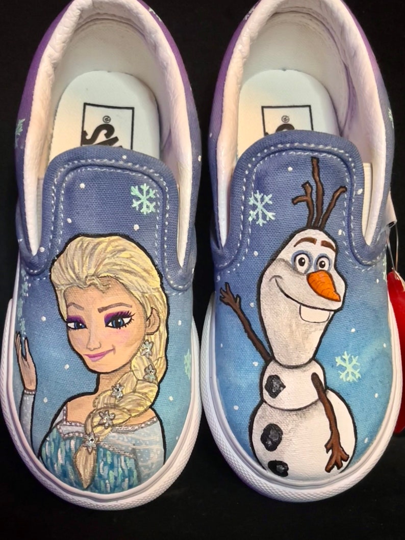 Puede incluir: Un par de zapatos de lona azul de deslizamiento con un dise&ntilde;o pintado a mano de Elsa y Olaf de la pel&iacute;cula Frozen. Los zapatos tienen suelas blancas y una franja blanca alrededor de la parte superior.