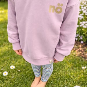 Könnte beinhalten: Ein lilafarbener Sweatshirt mit dem Wort "nö" in goldfarbenen Buchstaben. Das Sweatshirt wird mit gestreiften Leggings in Blau-, Grün- und Lilatönen kombiniert. Die Person steht auf grünem Gras mit Gänseblümchen.