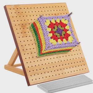 May include: A wooden crochet blocking board with a stack of colorful crochet squares pinned in place. The board is made of light-colored wood and has numerous small holes. A set of metal pins is included.