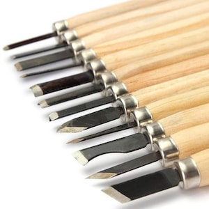 May include: A set of twelve wood carving tools arranged in a row, each featuring a sharp metal blade of varying shapes and sizes, a silver-toned metal ferrule, and a smooth, light-colored wooden handle, all aligned diagonally on a white background.
