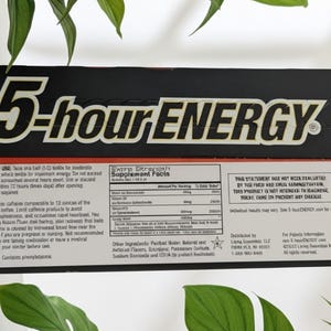 May include: A black and red 5-hour ENERGY box with white text. The product name is prominently displayed. The box contains supplement facts and usage instructions. The background includes green leaves.