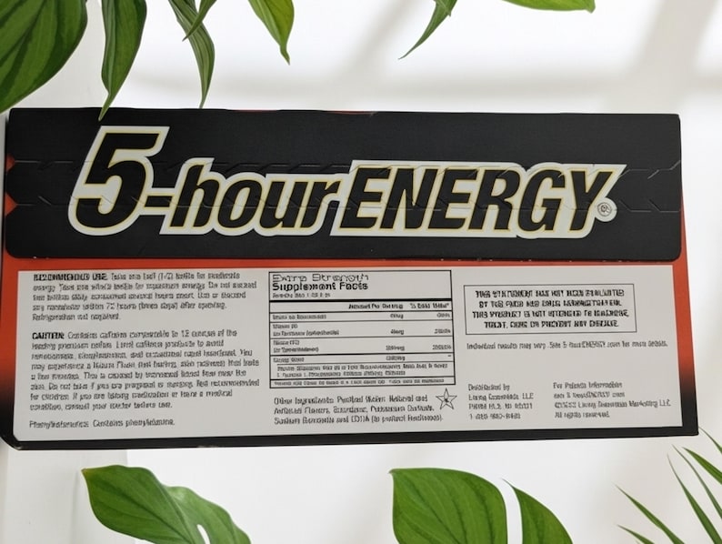 May include: A black and red 5-hour ENERGY box with white text. The product name is prominently displayed. The box contains supplement facts and usage instructions. The background includes green leaves.