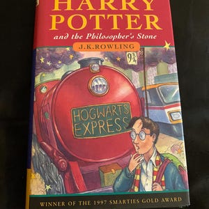 May include: A copy of "Harry Potter and the Philosopher's Stone" by J.K. Rowling. The book's cover is predominantly red, featuring a steam train with "Hogwarts Express" on it, and an illustration of a young boy. It is a winner of the 1997 Smarties Gold Award.