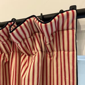 Puede incluir: Paneles de cortinas a rayas rojas y crema con ribete azul oscuro. Las cortinas cuelgan de una barra negra, creando un estilo clásico de granja. La tela tiene un patrón de rayas verticales.