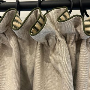 Peut inclure: Gros plan sur des rideaux en lin beige avec un dessus froncé. Les rideaux sont suspendus à une tringle noire avec des anneaux noirs. Le bord supérieur est bordé d'un tissu rayé vert et beige.