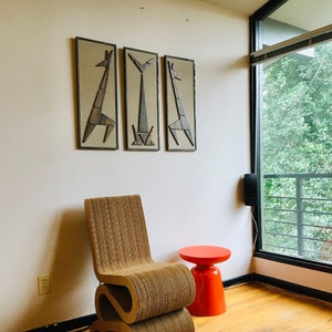 May include: A brown cardboard chair with a unique S-shaped design sits in a room with a red stool and three framed wall art pieces featuring geometric animal silhouettes.