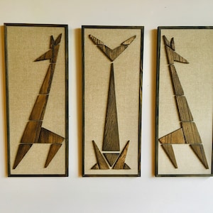 May include: Three framed wall art pieces featuring abstract geometric giraffe silhouettes made of wood, each with a different pose, on a beige linen background.