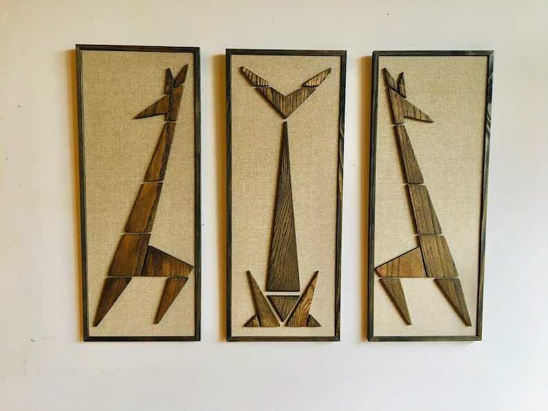 May include: Three framed wall art pieces featuring abstract geometric giraffe silhouettes made of wood, each with a different pose, on a beige linen background.
