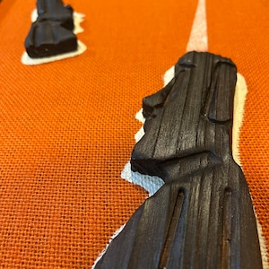 May include: Close-up of dark brown, carved Moai statues on a textured orange surface. The statues are on a white base. The image shows several statues in various stages of completion, with detailed facial features.