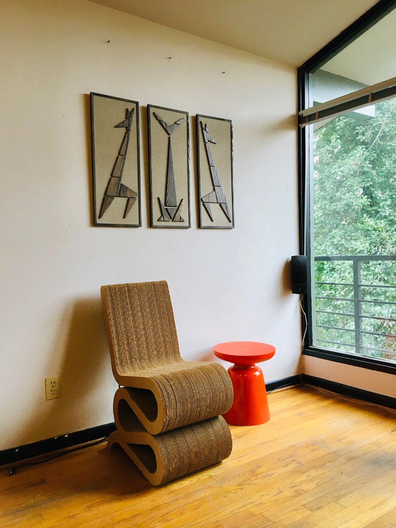 May include: A brown cardboard chair with a unique S-shaped design sits in a room with a red stool and three framed wall art pieces featuring geometric animal silhouettes.