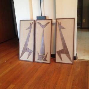 May include: Three framed wall art pieces with a geometric design of a giraffe. The frames are made of wood and have a brown finish. The background is a wooden floor.