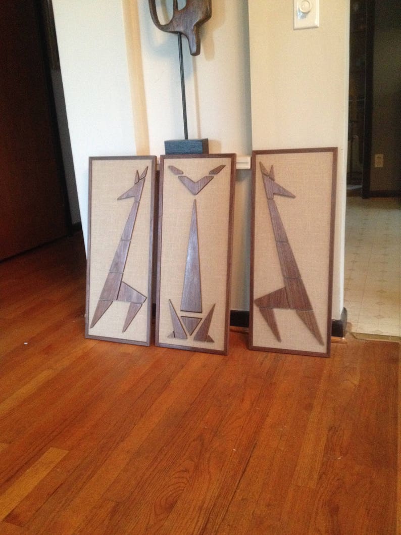May include: Three framed wall art pieces with a geometric design of a giraffe. The frames are made of wood and have a brown finish. The background is a wooden floor.