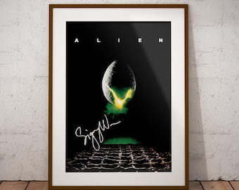 May include: A framed movie poster for Alien featuring a central cracked egg emitting a bright yellow and green glow against a black background. The word ALIEN is printed in white at the top, and a white signature is visible below the egg.