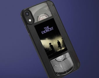 May include: A black smartphone case featuring a graphic design that mimics the appearance of a vintage VHS cassette tape. The center label displays the movie title The Exorcist with an illustration of a figure standing under a street lamp.