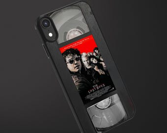 May include: A black smartphone case featuring a graphic design that mimics a VHS tape. The center displays the movie poster for The Lost Boys, showing characters in a red and black color scheme. The case includes clear reel windows at the top and bottom.