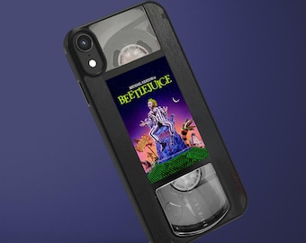 May include: A black smartphone case designed to resemble a vintage VHS cassette tape. The center features a colorful graphic print of the Beetlejuice movie poster, including the text Michael Keaton in Beetlejuice, with clear reel windows.