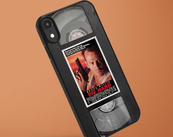 May include: A black smartphone case designed to resemble a vintage VHS cassette tape. The center features the Die Hard movie poster with Bruce Willis holding a gun, while the top and bottom sections display clear plastic reel windows.