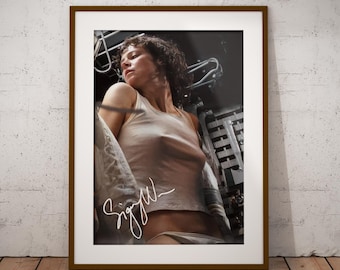 May include: A framed photographic print featuring a person with curly dark hair wearing a white tank top and white underwear. The image includes a white signature across the lower portion of the print. The frame is a solid brown color.