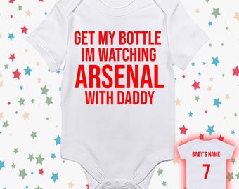 May include: White baby bodysuit with red text that reads "GET MY BOTTLE IM WATCHING ARSENAL WITH DADDY." A smaller bodysuit shows "BABY'S NAME" and the number "7" in red. The background has a pattern of colorful stars.