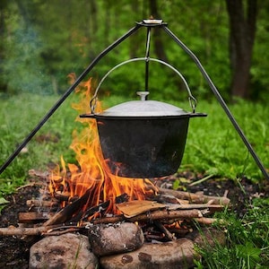 Peut inclure: Une marmite noire avec un couvercle argenté est suspendue au-dessus d'un feu de camp. Le pot est suspendu à un trépied en métal noir. Des flammes et de la fumée s'élèvent du feu, avec de l'herbe verte et des arbres en arrière-plan.