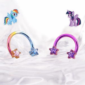 May include: Two horseshoe-shaped body jewelry pieces. One is rainbow-colored with a gold star on each end and a blue star in the middle. The other is purple with purple star accents. Cartoon pony stickers are in the background.