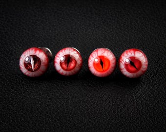 May include: Four round stud earrings featuring a realistic red eye design with a vertical black pupil. Each earring has a white sclera with detailed red blood vessels radiating outward, creating a distinct ocular appearance for each piece.