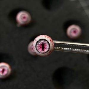 Könnte beinhalten: Nahaufnahme mehrerer kleiner, runder Glasaugenkomponenten. Die Augen haben eine weiße Iris mit rosa und roten Adern und eine vertikale, dunkelrosa Pupille. Ein Auge wird von einer Pinzette gehalten. Der Hintergrund ist dunkel.