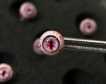 May include: A spherical glass eye cabochon held by metal tweezers, featuring a vertical black pupil surrounded by a vibrant pink iris with intricate red vascular detailing on the white sclera, set against a dark foam background with circular cutouts.