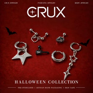 May include: A collection of silver-toned body jewelry pieces including a heart charm barbell, a skull stud, a star hoop earring, a black guitar barbell, a star-patterned hoop, and a dangling star charm earring, with text reading CRUX HALLOWEEN COLLECTION.