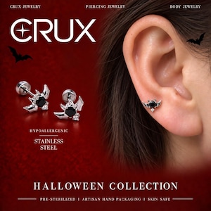 May include: A pair of silver-toned bat-shaped stud earrings featuring a central round black gemstone and a small clear accent stone. The text CRUX JEWELRY, PIERCING JEWELRY, BODY JEWELRY, HYPOALLERGENIC STAINLESS STEEL, and HALLOWEEN COLLECTION is visible.