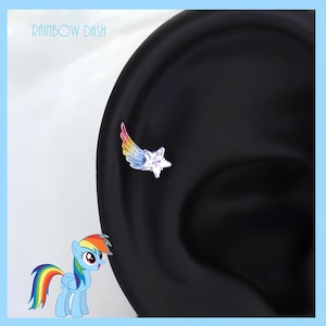 May include: A silver earring featuring a star and a rainbow-colored wing design. The earring is set against a black background, with the text "Rainbow Dash" at the top. A cartoon pony is in the lower left corner.