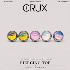 May include: Five circular piercing tops from Crux Jewelry arranged in a horizontal row. Each piece features a smooth, iridescent cabochon stone set in a metallic frame. The text reads Crux Jewelry, Piercing Jewelry, Replacement End, Pre-Sterilized, Artisan Hand Packaging, Skin Safe, Piercing Top, and Body Jewelry.