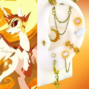 May include: A collection of gold-toned ear piercings displayed on a white ear model, featuring a hand charm, double chains, a sun-shaped stud with a pink center, a white wing, a cross, and a dangling star-shaped charm with a red accent.