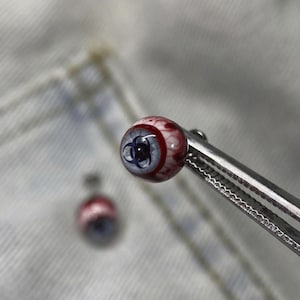 Könnte beinhalten: Nahaufnahme einer kleinen, runden, augapfelförmigen Perle, die von einem Metallwerkzeug gehalten wird. Die Perle hat ein rot-weißes Muster mit einer blauen Iris und einem Biohazard-Symbol. Eine weitere Perle ist im Hintergrund unscharf.