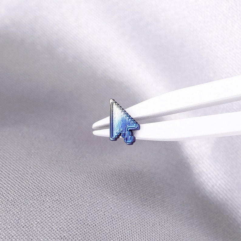May include: A small, metallic computer mouse cursor icon with a gradient finish transitioning from white to deep blue. The cursor is held by white plastic tweezers, showcasing its sharp, pixelated edges and distinct arrow shape in a compact size.