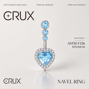Pode incluir: Um piercing de umbigo em titânio ASTM F136 da marca CRUX com uma pedra central azul-clara em forma de coração rodeada por um halo de pequenas pedras transparentes. A haste vertical inclui três pedras azul-claras alinhadas.