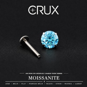 May include: A round, brilliant-cut blue moissanite stone set in a four-prong metal stud earring with a flat-back post. The text CRUX, MOISSANITE, ONE STONE ONE CERTIFICATE, DIAMOND TESTER VERIFIED, and piercing locations are visible.