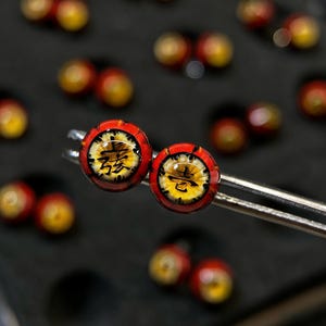 以下が含まれることがあります： 中国語の文字が入った赤と黄色の丸いイヤリング2つのクローズアップ。 イヤリングは黒い縁取りがあり、銀色のツールで保持されています。 他の同様のイヤリングが背景にぼやけています。